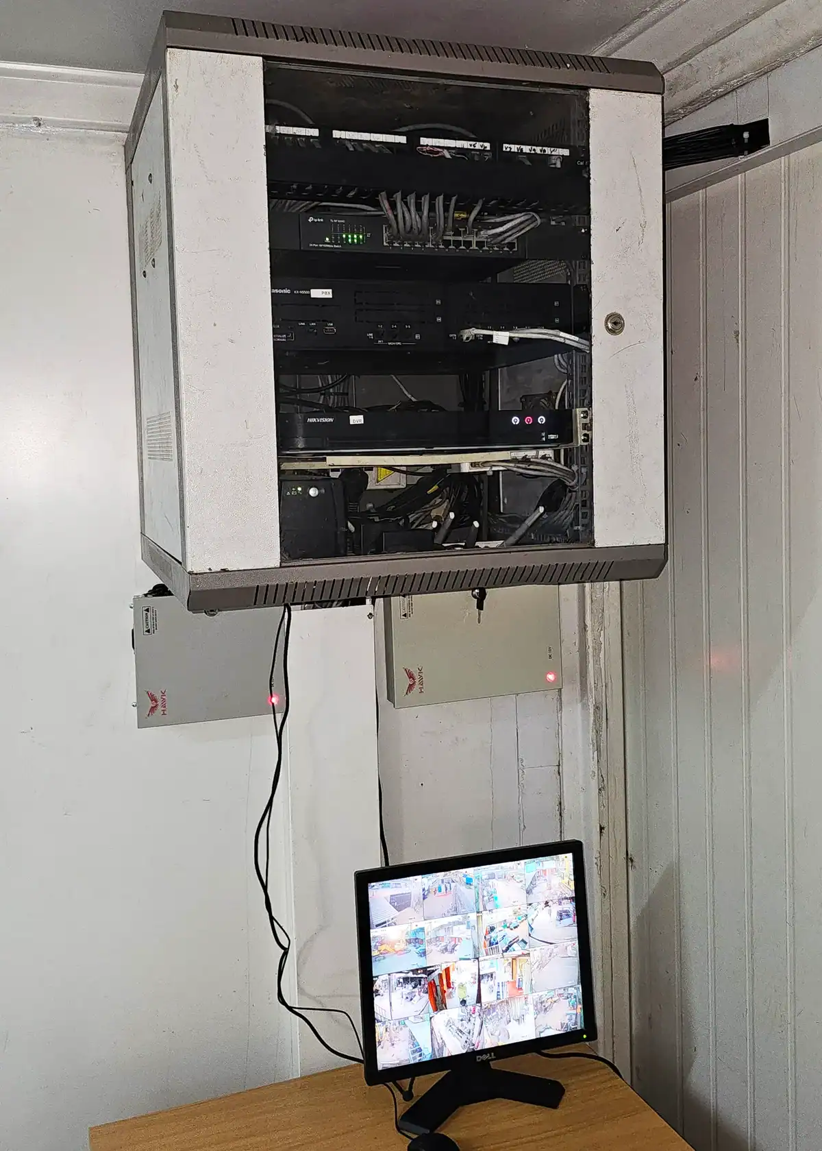 Wall-mounted network rack with surveillance equipment and CCTV monitoring screen displaying multiple camera feeds.